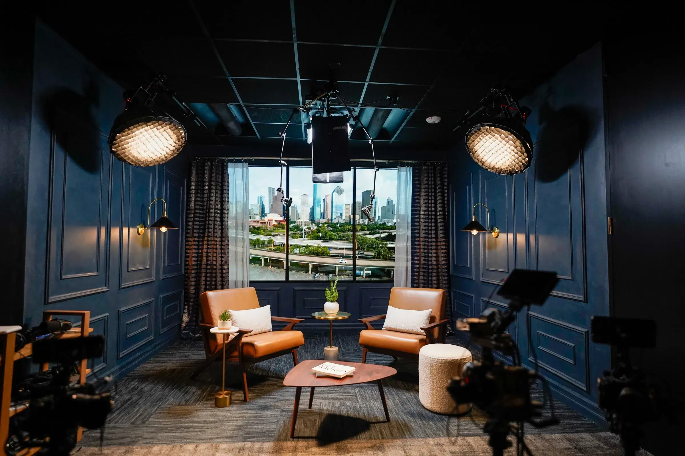 The finished Anthem interview studio — navy paneled walls, tan leather chairs facing a wide window onto the Houston skyline, cameras and overhead film lighting rigged around the set