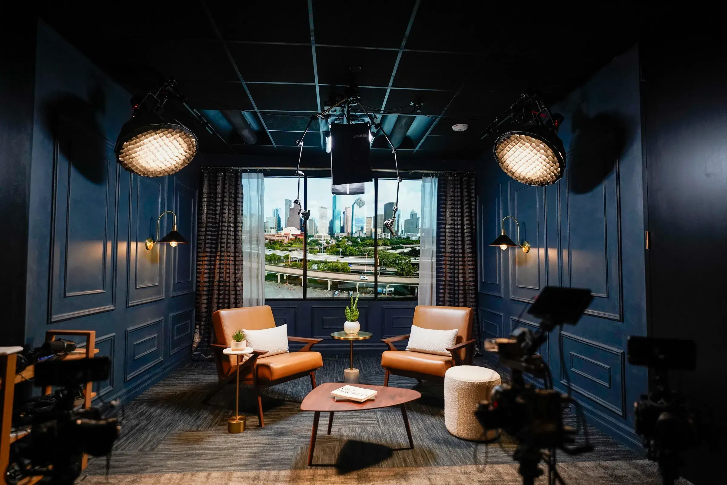 The finished Anthem interview studio — navy paneled walls, tan leather chairs facing a wide window onto the Houston skyline, cameras and overhead film lighting rigged around the set