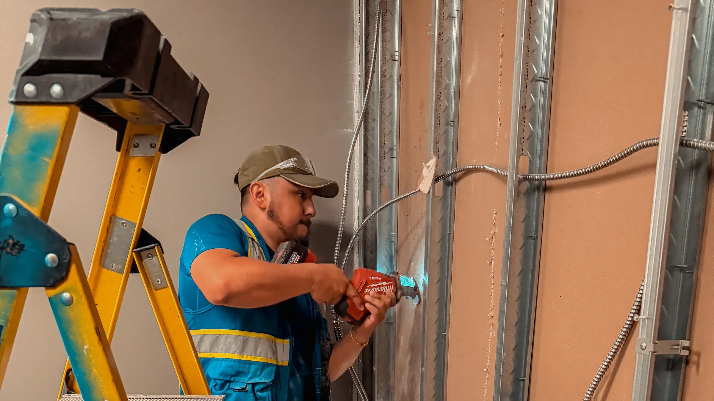 An electrician on a ladder drilling into metal stud framing, running armored cable to boxes set in the open wall