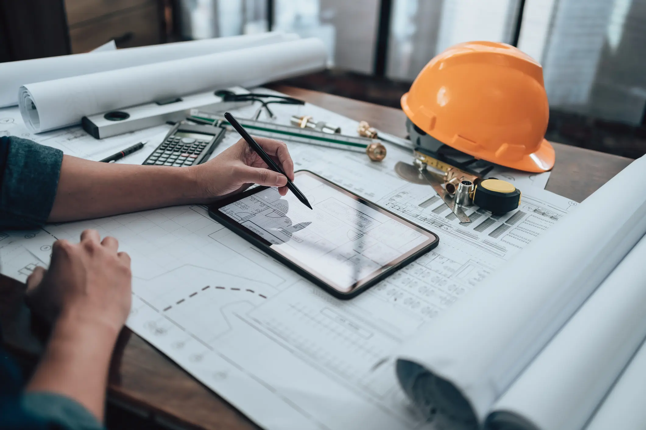 An estimator pricing a project on a tablet over blueprints, beside a hard hat and calculator