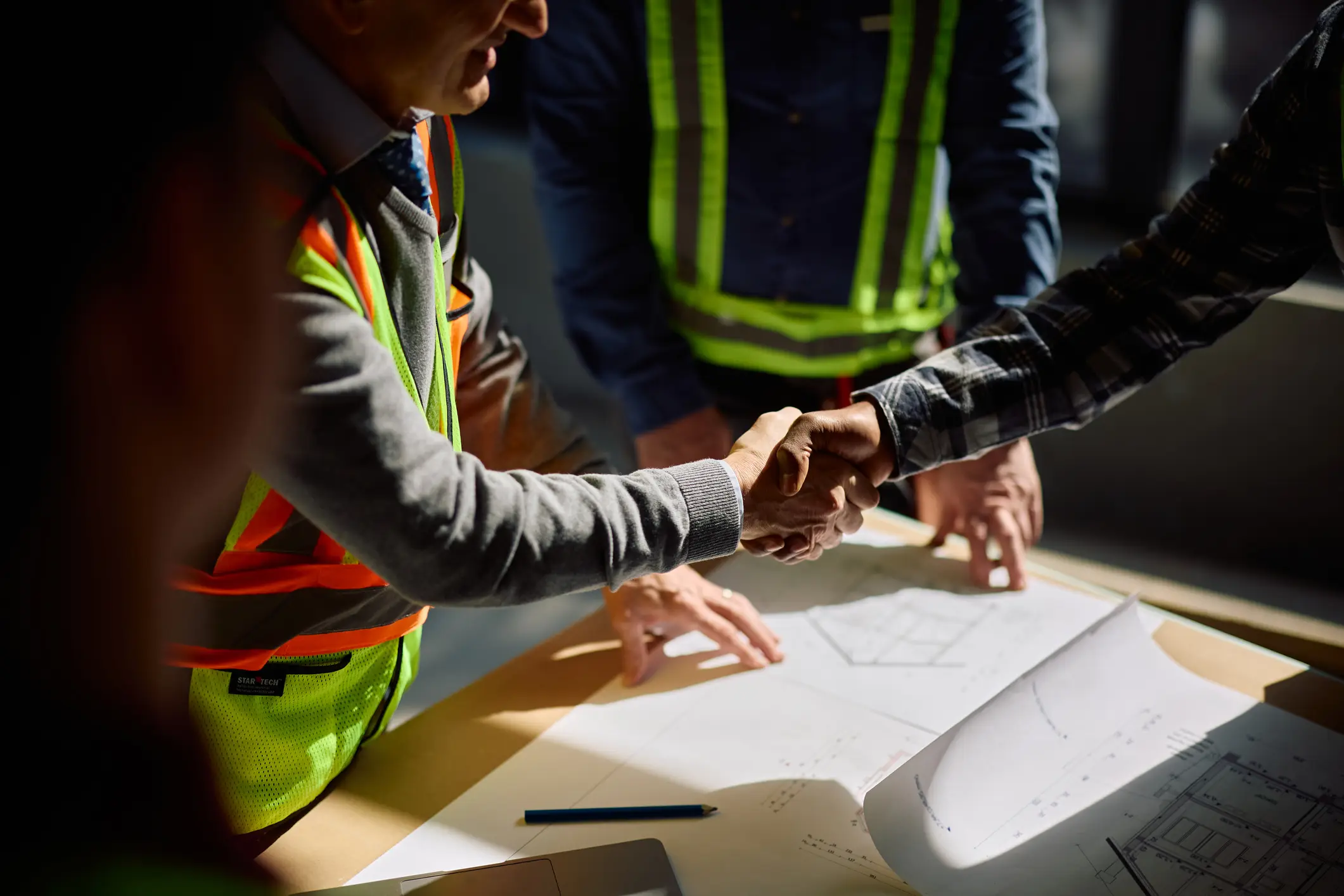 A contractor and an owner shaking hands over blueprints at a construction site