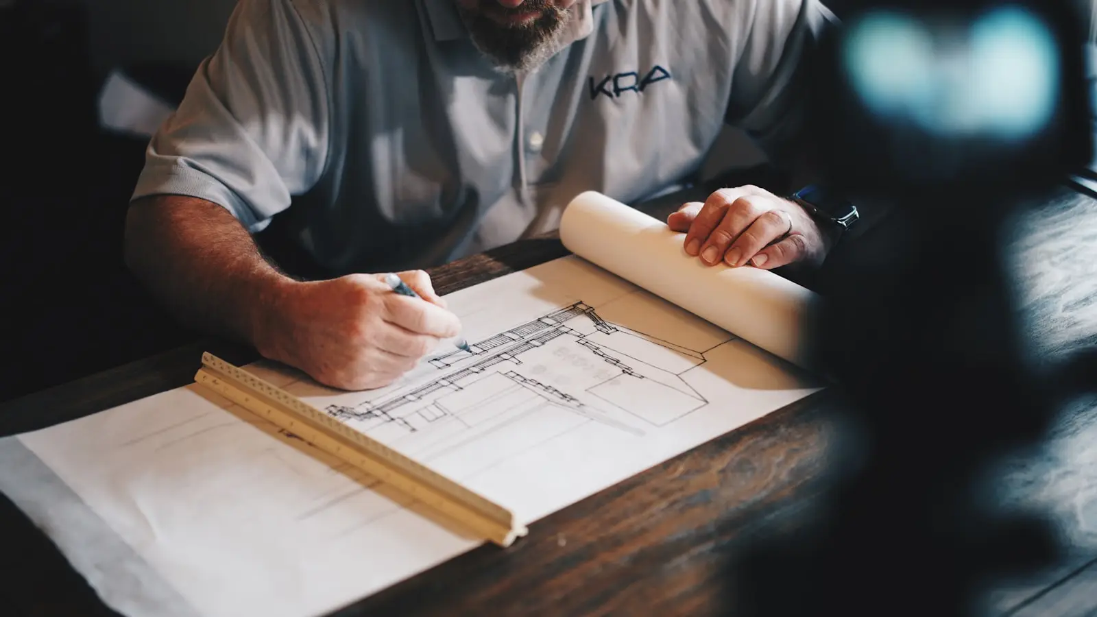 A builder sketching a floor plan over blueprints with a ruler at a wood desk