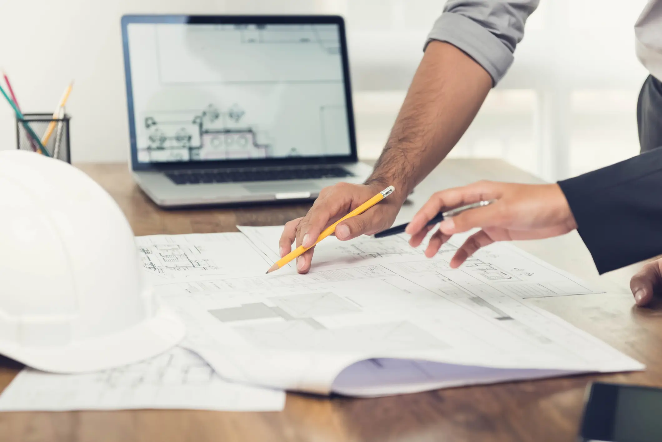 A contractor and client reviewing construction blueprints over a desk with a hard hat and a laptop showing plans