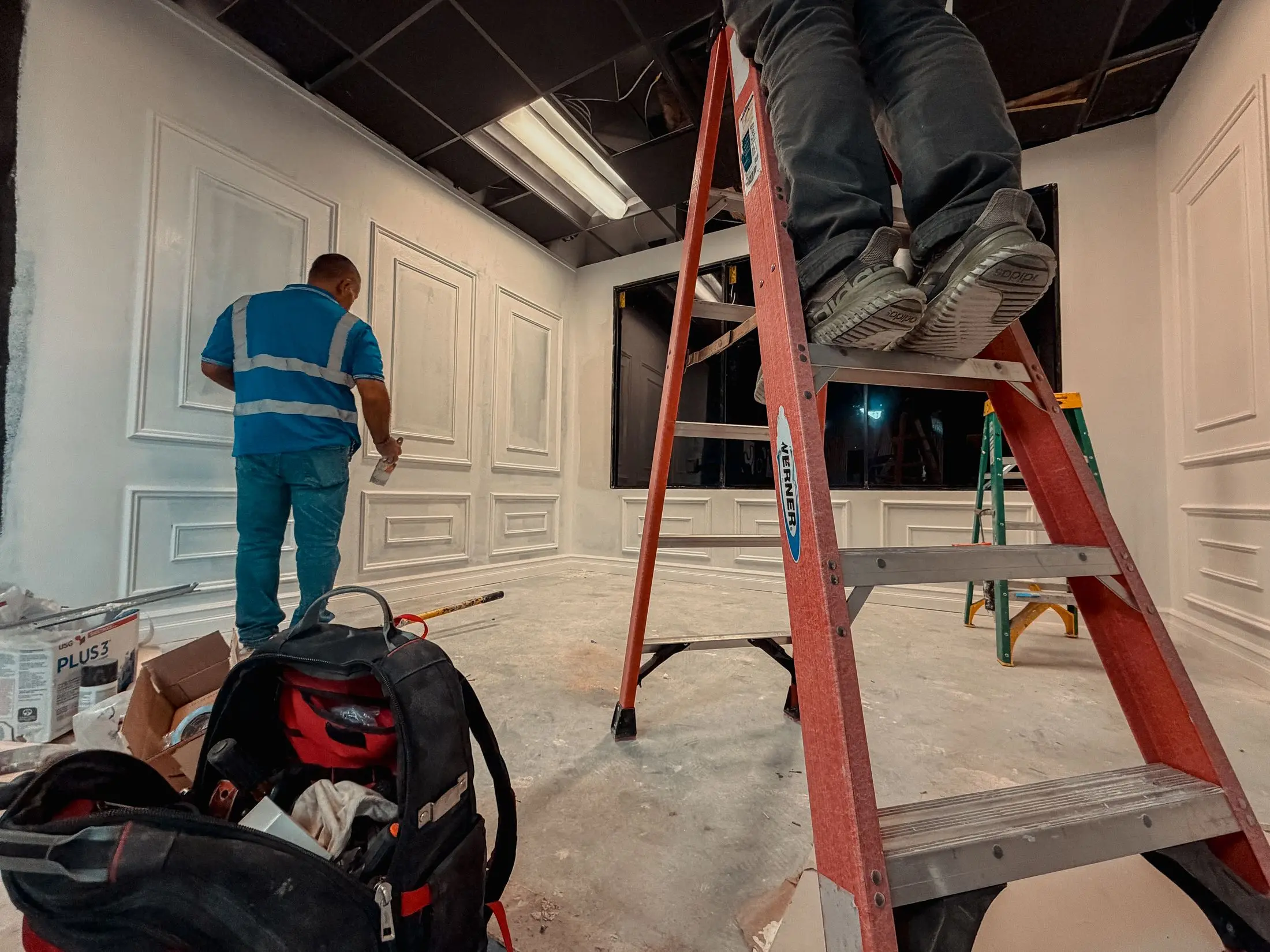 Two workers finishing an interior build-out — one taping wall panels, another on a ladder working in the open ceiling