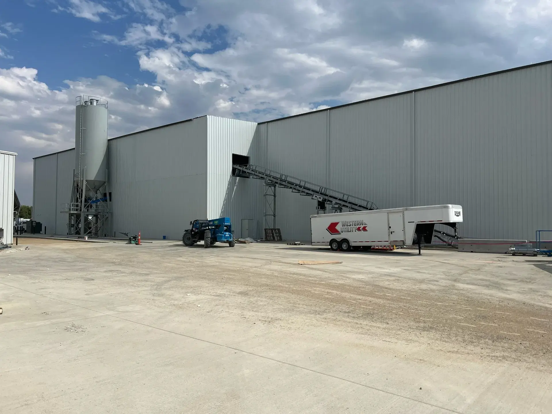 The batch plant silo and conveyor running into the side of the production building, with a telehandler and materials trailer on the new concrete apron