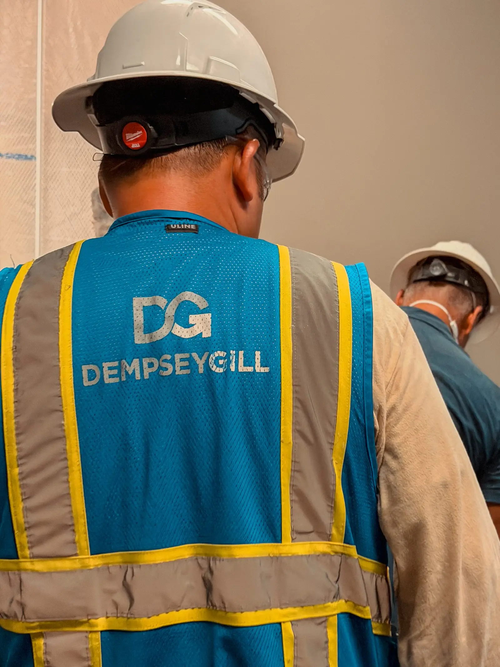 A DempseyGill field crew member in a branded safety vest and hard hat working alongside a subcontractor on an interior build-out