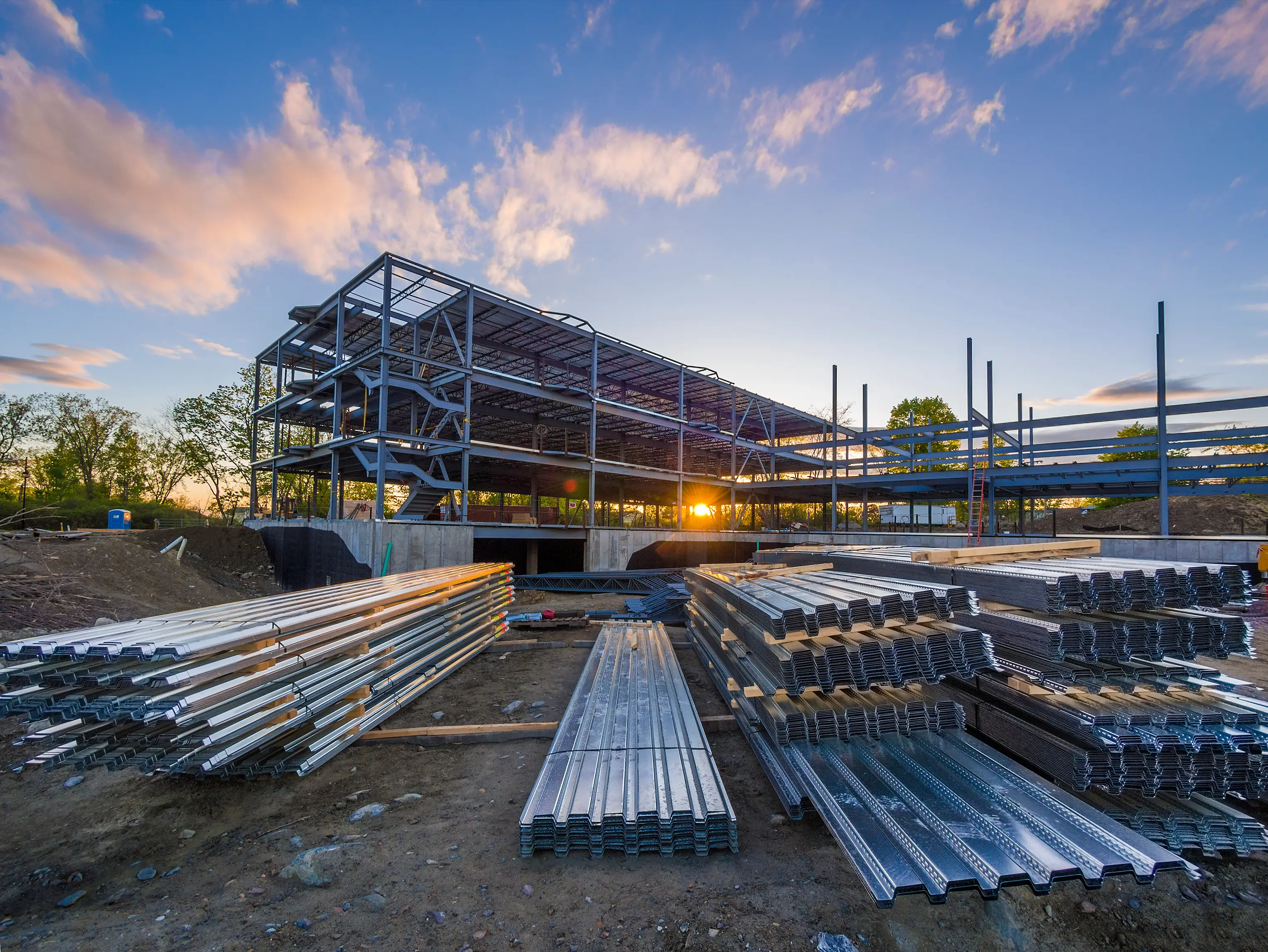 A steel-frame commercial building under construction at sunset, with staged steel decking in the foreground