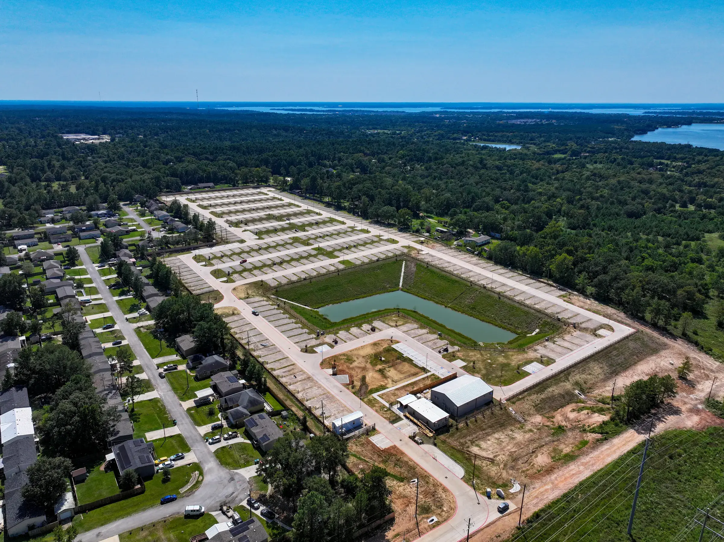 Aerial view of the Willis Lakes development — rows of concrete pads on paved, lit streets around a lined detention pond, with Lake Conroe on the horizon
